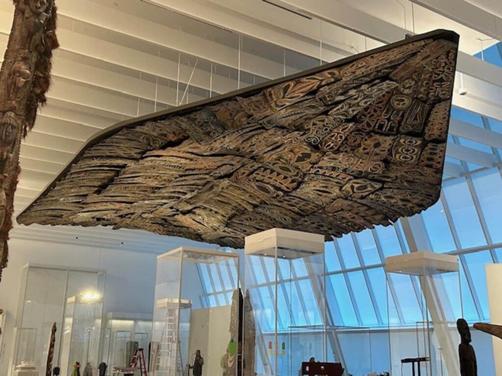 A large, ornately carved wooden canoe is suspended from the ceiling in a spacious, modern museum gallery with white walls and large windows. Several display cases and various artifacts are visible below.