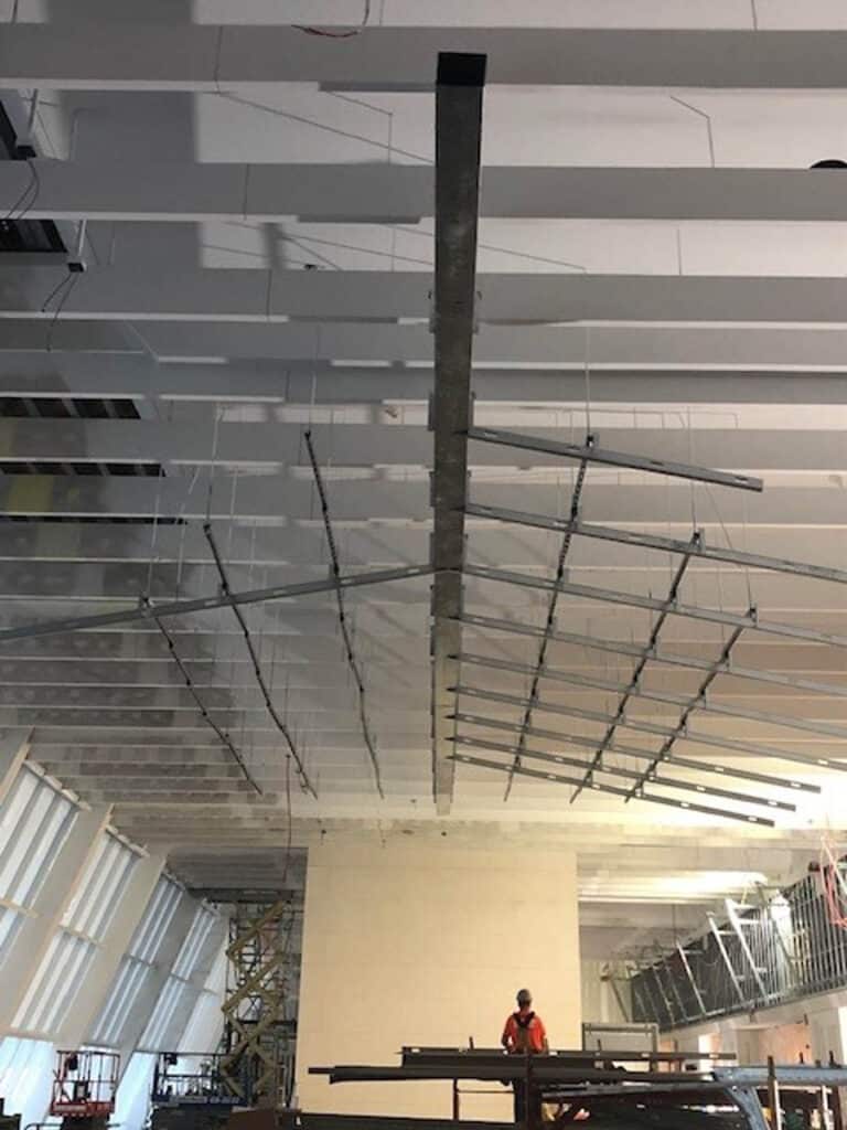 A construction worker stands below a partially built ceiling with misaligned beams. The slanted windows on the left and construction scaffolding indicate the building is under construction.
