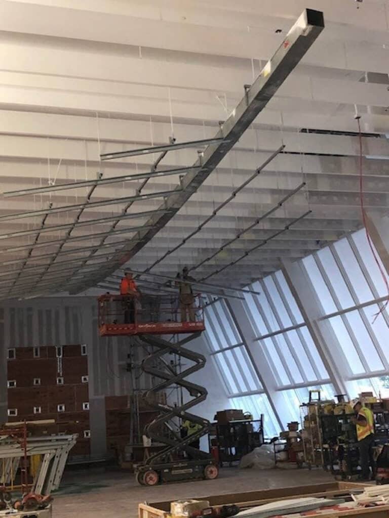 Two construction workers on a scissor lift work on a partially installed metal framework suspended from a high, slanted ceiling in an unfinished building. Other workers and construction materials are visible below.