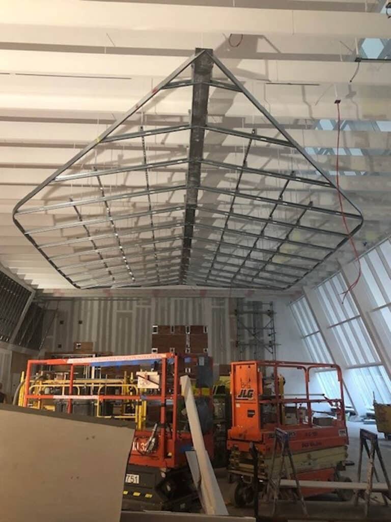 A large metal frame shaped like a paper airplane hangs from the ceiling of a construction site, with scaffolding, lifts, and building materials scattered below.