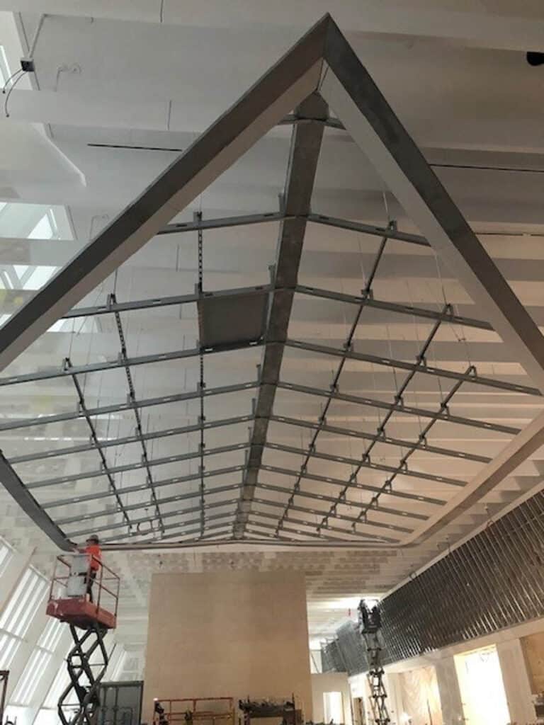 A large triangular metal frame is suspended from a high ceiling in a spacious building under construction. Workers on lifts are installing or inspecting the structure. Scaffolding and construction materials are visible below.