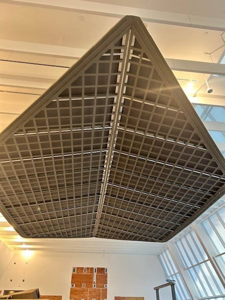 Large suspended grid structure hanging from a ceiling in a modern interior space with white beams, windows, and partial brick walls visible in the background.