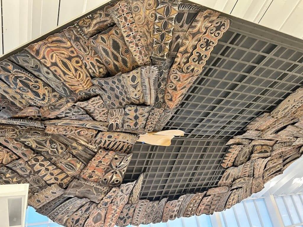Decorative wooden panels with intricate, tribal-inspired carvings and earthy colors are arranged in a circular pattern on a ceiling, surrounding a central grid structure. Light filters in from windows nearby.