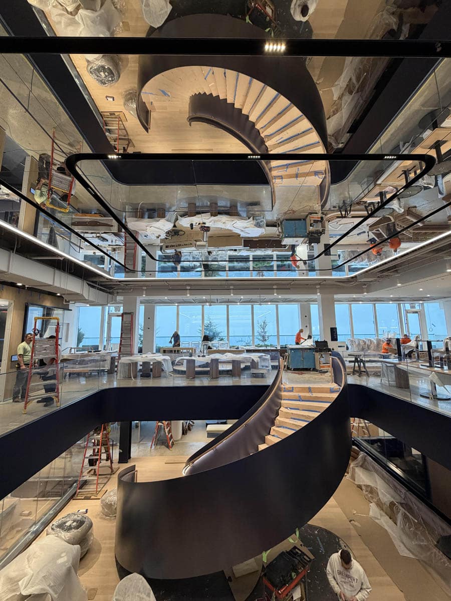 A modern interior features a dramatic spiral staircase with black railings, wooden steps, and a mirrored ceiling that reflects the staircase. Workers are present, suggesting ongoing construction or setup. Large windows offer outdoor views.