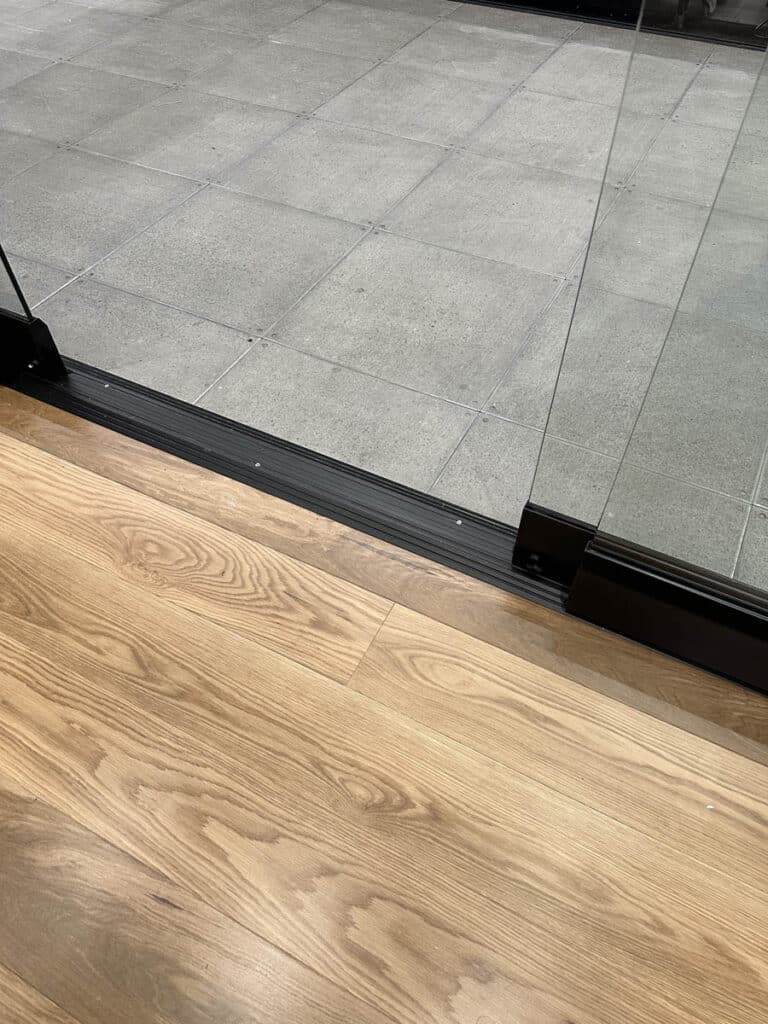 A glass door separates a wooden floor indoors from a gray tiled outdoor area. The door has a black metal frame at the bottom and is partially open.