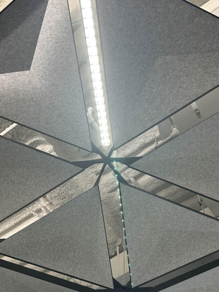 Ceiling view with triangular gray acoustic panels arranged in a geometric pattern, reflecting a long fluorescent light fixture and exposed ductwork above.