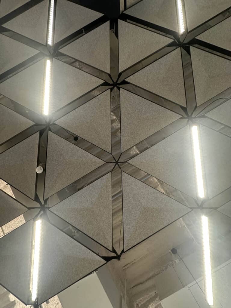 Geometric ceiling design with triangular panels forming hexagonal patterns, intersected by bright linear LED lights. The panels are gray, and parts of exposed ceiling and pipes are visible in the background.