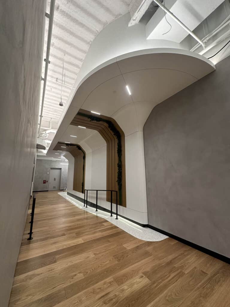 A modern hallway with wooden floors, curved white ceilings, and sleek architectural details, featuring recessed lighting, a light gray wall, and a black handrail along a raised platform.