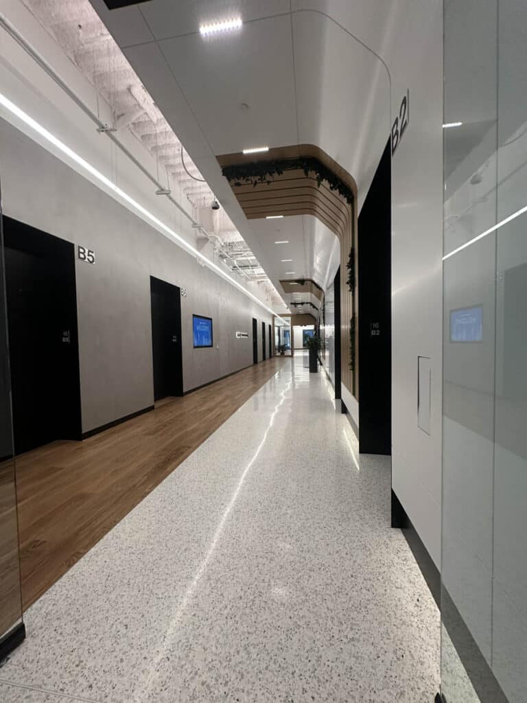 A modern office hallway with polished terrazzo floors, wood accents, gray walls, and black doorways labeled with signs like 