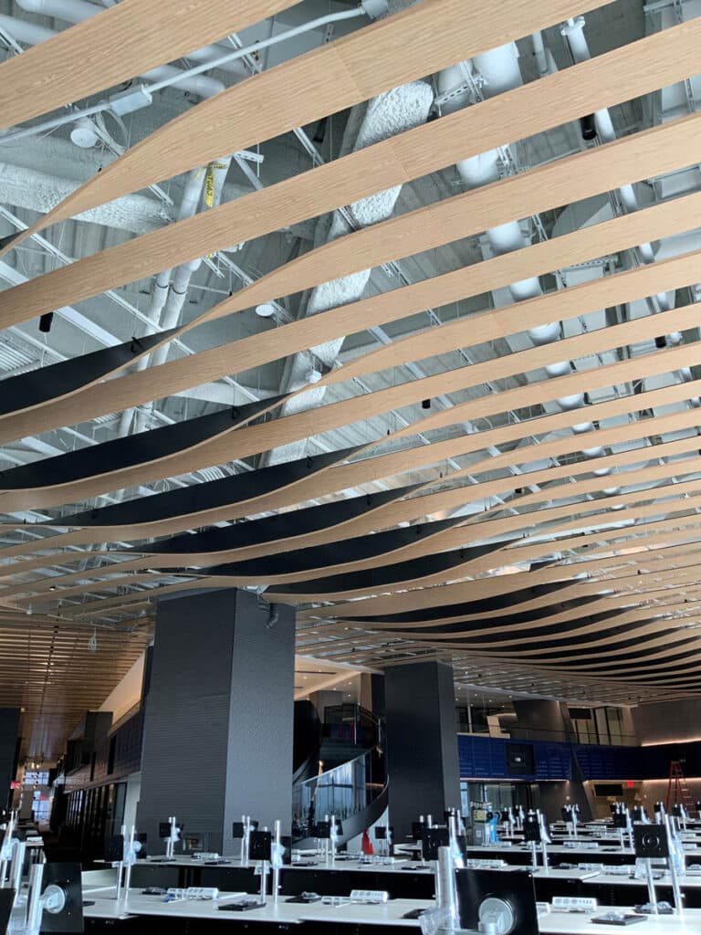 Curved wooden slats form an artistic wave pattern on the ceiling of a modern, open indoor space with industrial fixtures, dark columns, and unfinished seating or tables beneath.