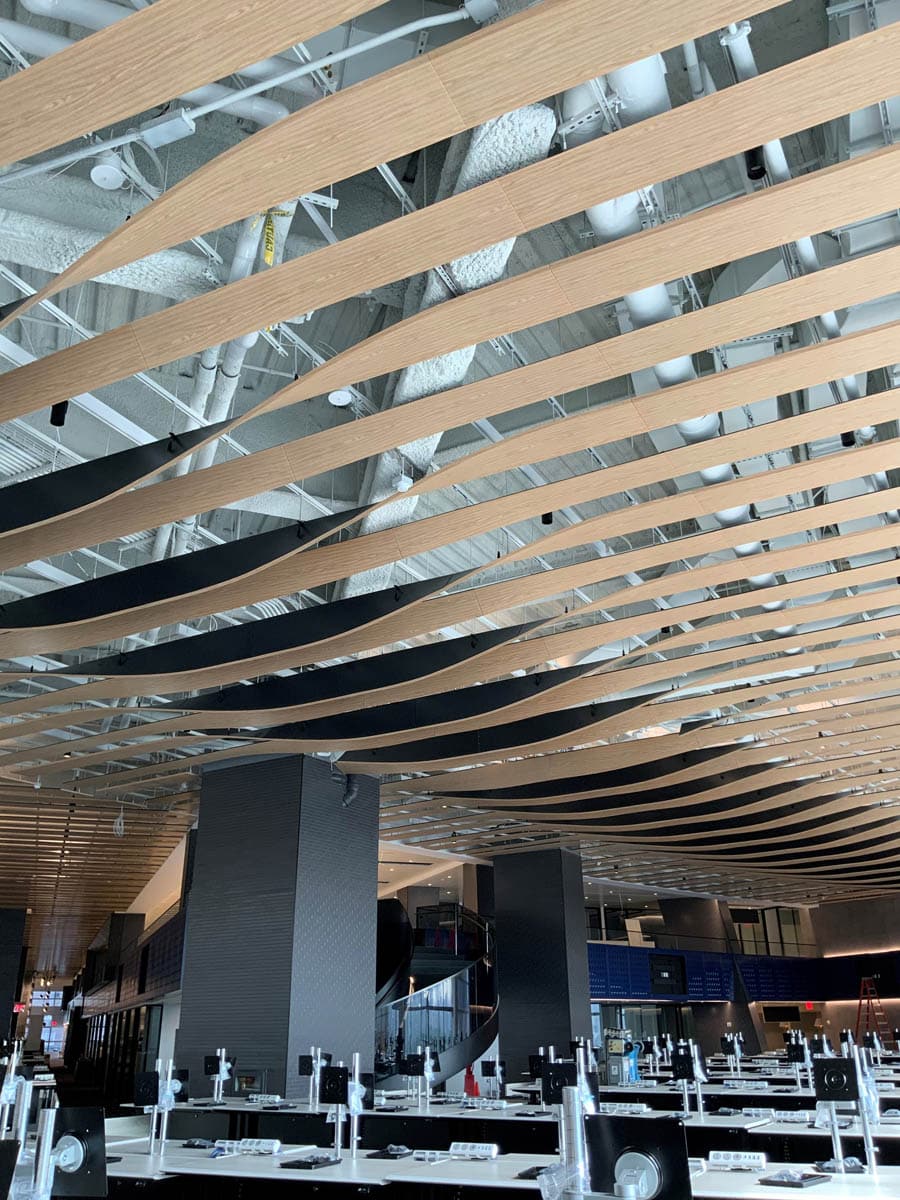 Curved wooden slats form an artistic wave pattern on the ceiling of a modern, open indoor space with industrial fixtures, dark columns, and unfinished seating or tables beneath.