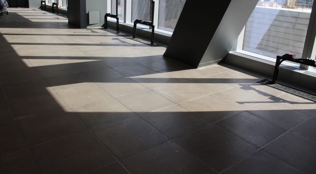 Sunlight casts large, angular shadows from structural columns onto a tiled floor inside a modern building with large windows and a view of nearby skyscrapers.