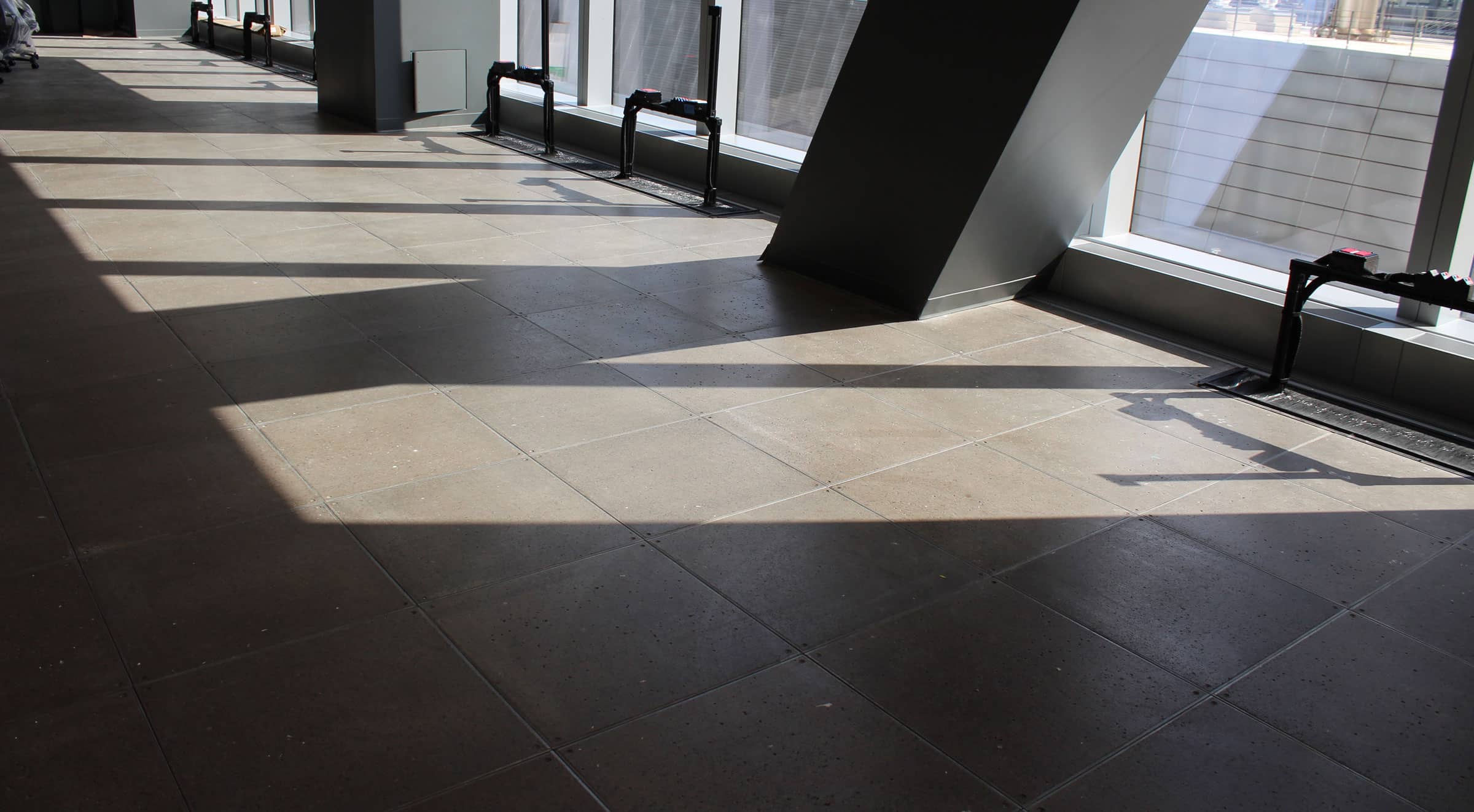 Sunlight casts large, angular shadows from structural columns onto a tiled floor inside a modern building with large windows and a view of nearby skyscrapers.