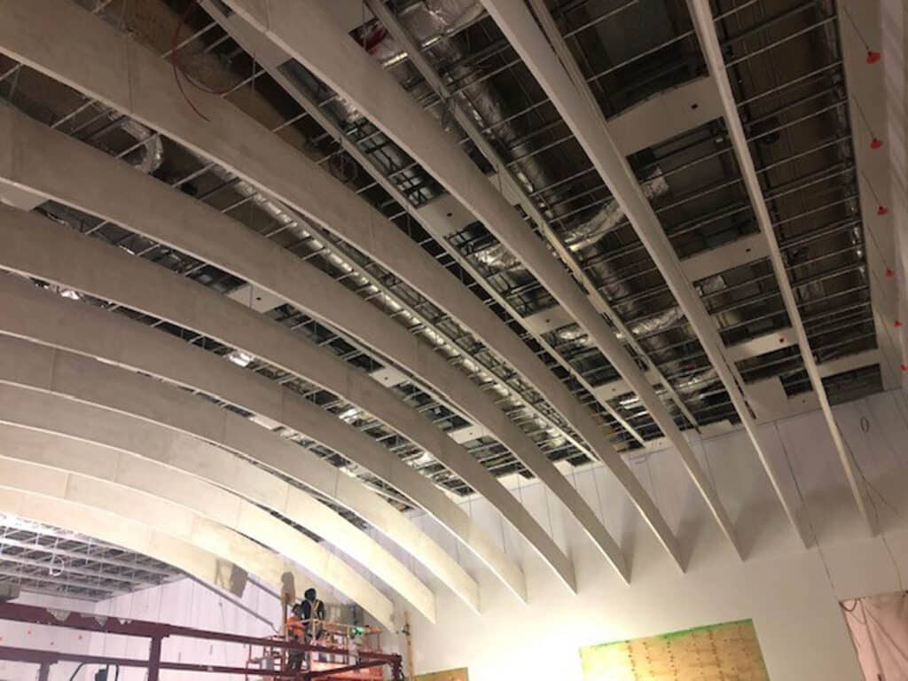 A partially constructed building interior with large curved beams on the ceiling, exposed ductwork, and workers on a lift performing installation or maintenance.