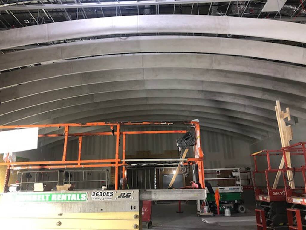 An indoor construction site with arched ceiling beams, scaffolding, construction equipment, and tools scattered around the unfinished area. Exposed beams and partially installed ceiling panels are visible.