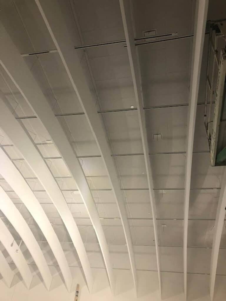 A ceiling with white curved beams and several horizontal glass panels installed between them. There is a single white ball resting on one of the glass panels.