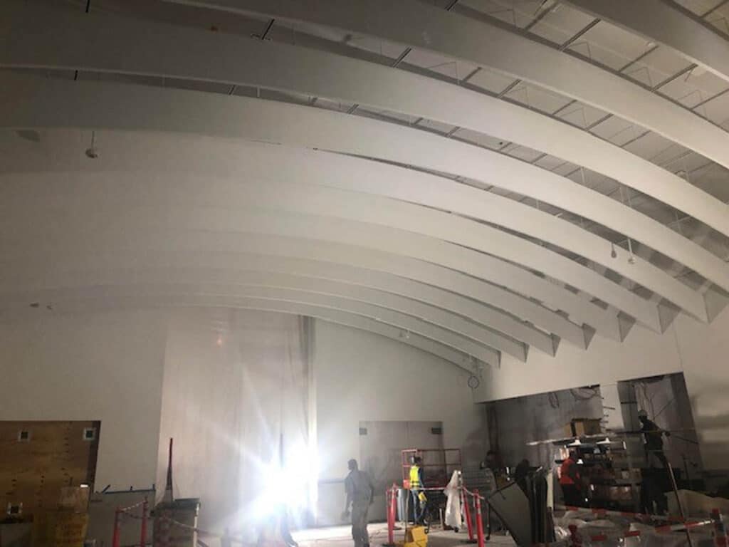 A large indoor space under construction with curved white ceiling beams, construction materials scattered on the floor, and workers in safety vests standing near a bright light source.