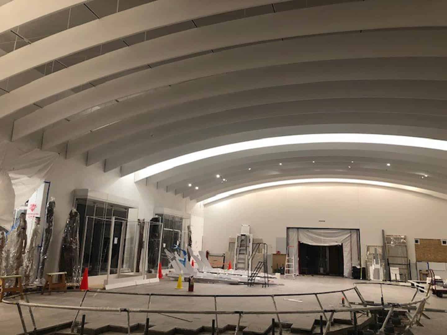 A large indoor space under construction features high curved ceilings, exposed beams, construction materials, scaffolding, traffic cones, and plastic-covered doorways. The area is unfinished, with tools and debris scattered.
