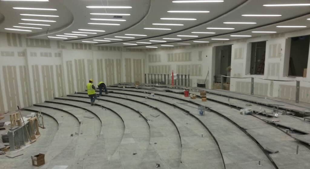 A large, modern lecture hall under construction, with curved, tiered seating and unfinished walls. Two workers in yellow safety vests stand in the center, surrounded by construction materials and equipment.