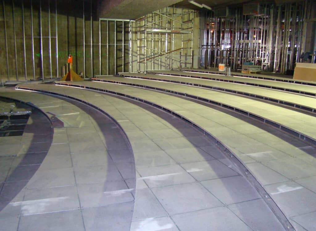 Rows of unfinished tiered seating in a construction site, with exposed metal framing on the walls, visible scaffolding, and construction materials scattered around.