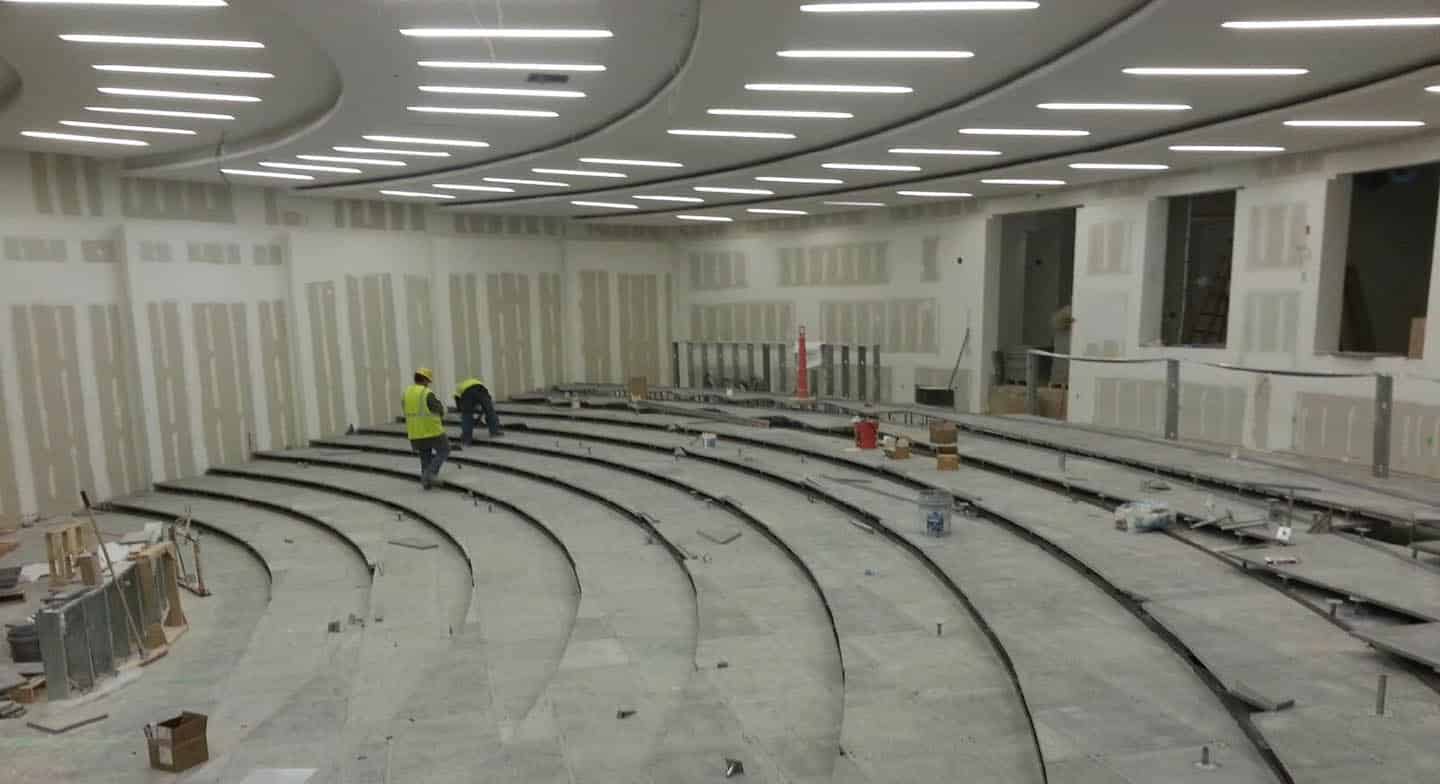 A large, modern lecture hall under construction, with curved, tiered seating and unfinished walls. Two workers in yellow safety vests stand in the center, surrounded by construction materials and equipment.