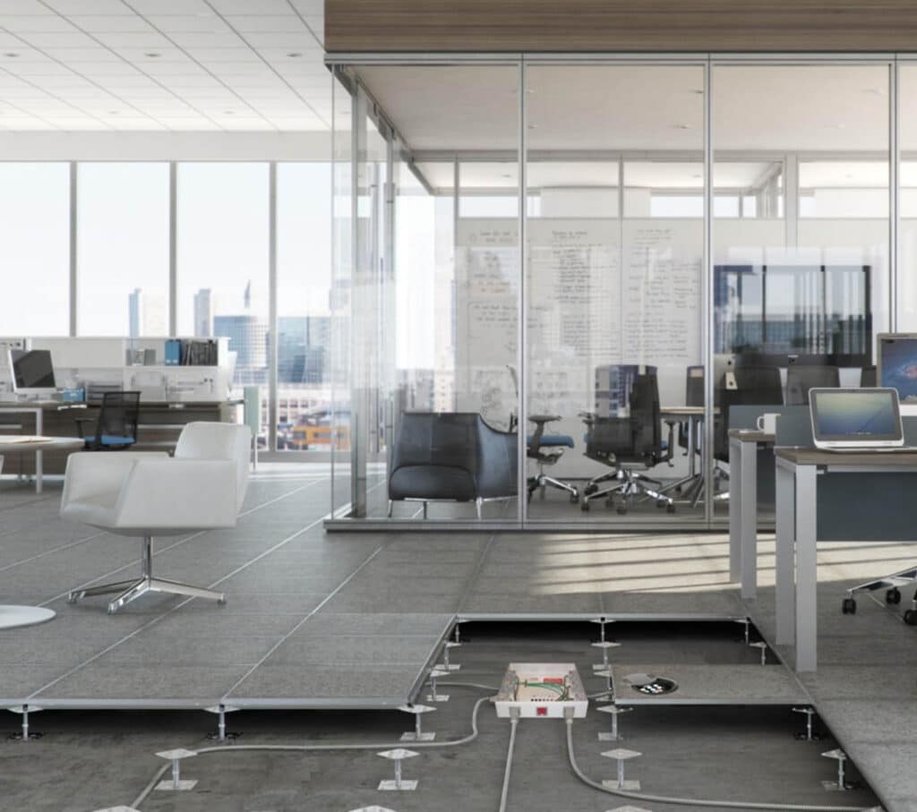 Modern office with glass-walled meeting room, desks, and chairs. Floor panels are lifted, revealing electrical wiring and cables underneath the raised flooring system. Cityscape visible through large windows.