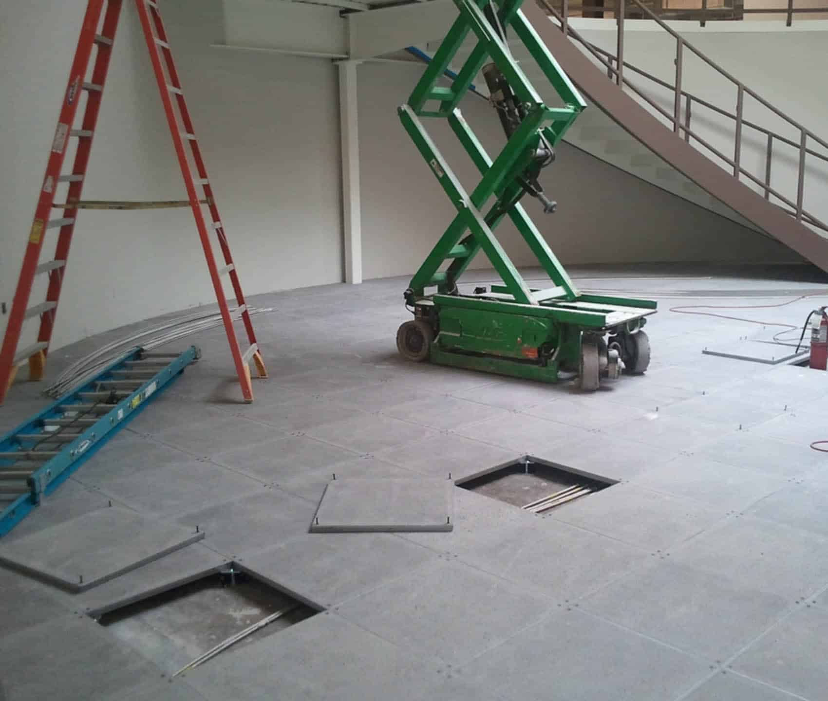 A room under construction with an open raised floor showing cables, a green scissor lift, two ladders (one red, one blue), and a staircase in the background. Some floor panels are removed.