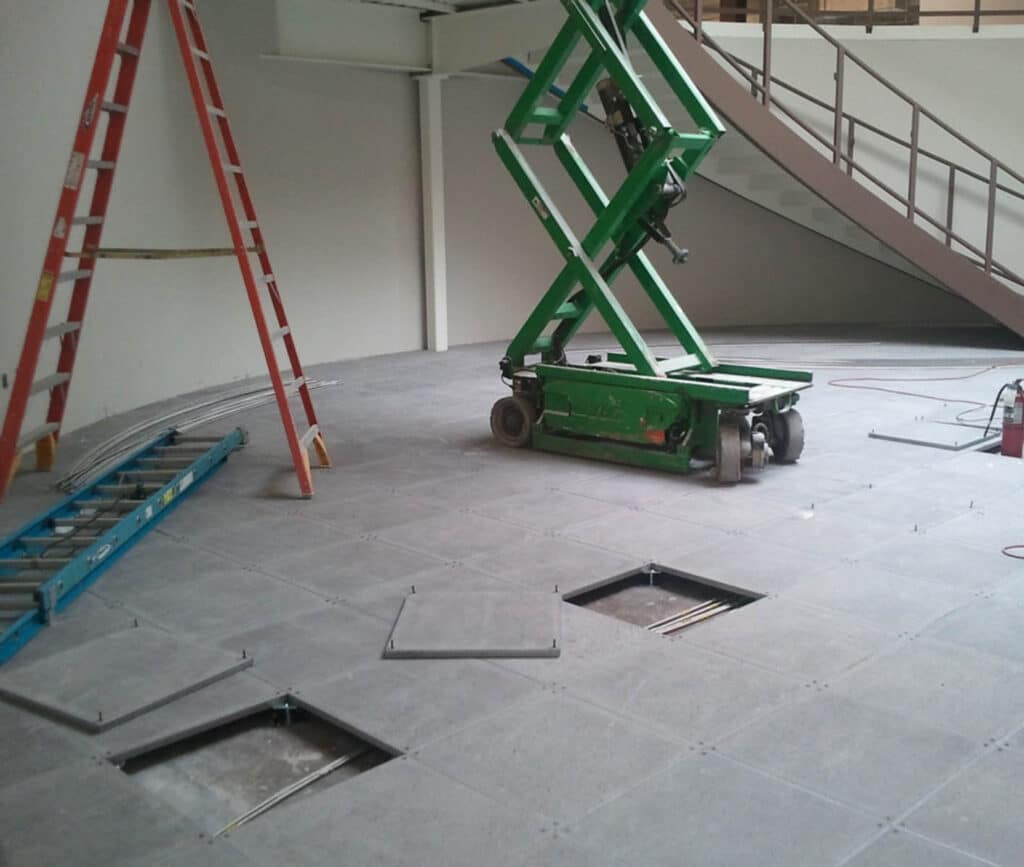 A room under construction with a green scissor lift, two ladders, and several raised floor tiles removed, exposing cables underneath. A staircase curves upward in the background.