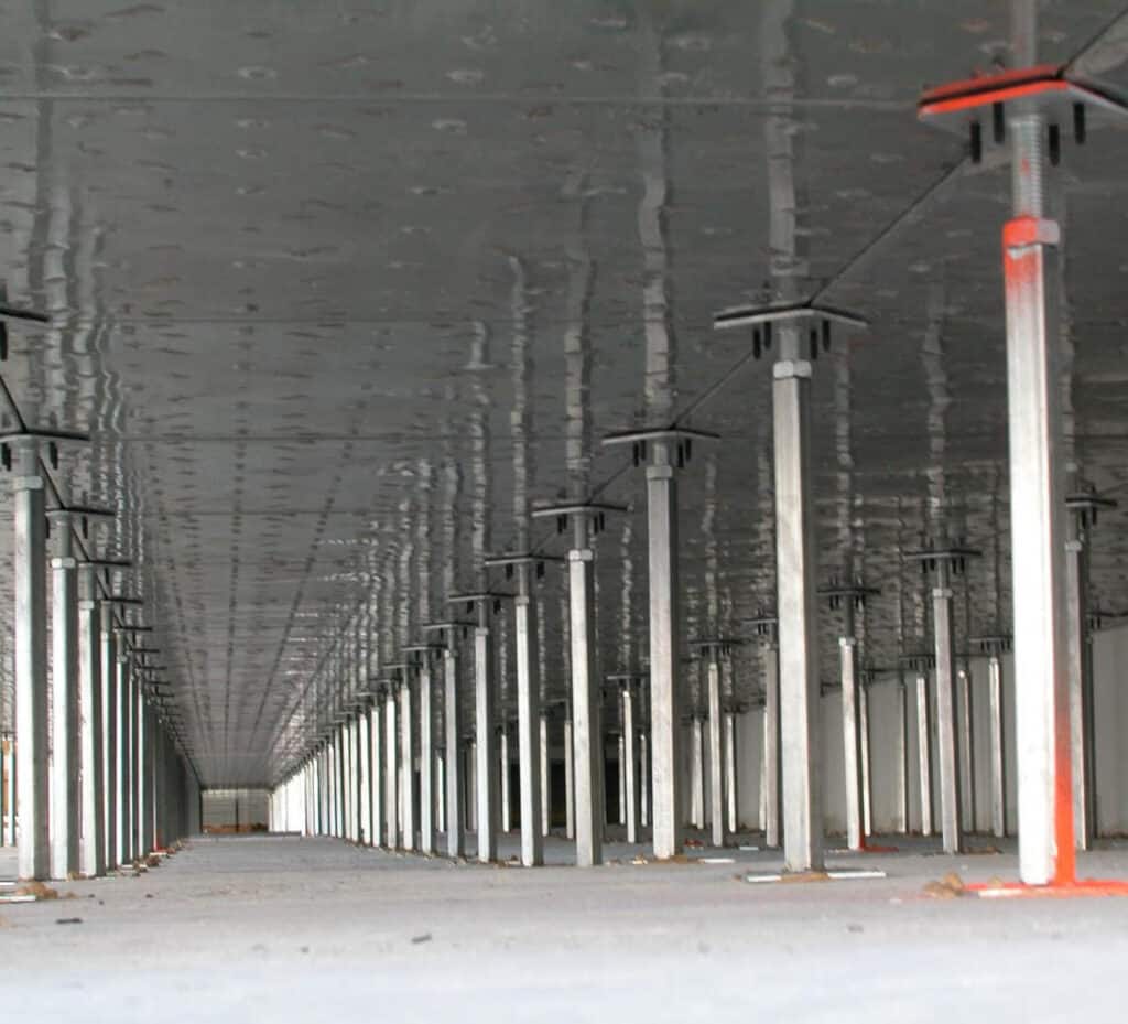 A view beneath a large concrete slab supported by numerous metal scaffolding poles, arranged in rows, providing temporary support during construction. The space is empty and industrial.